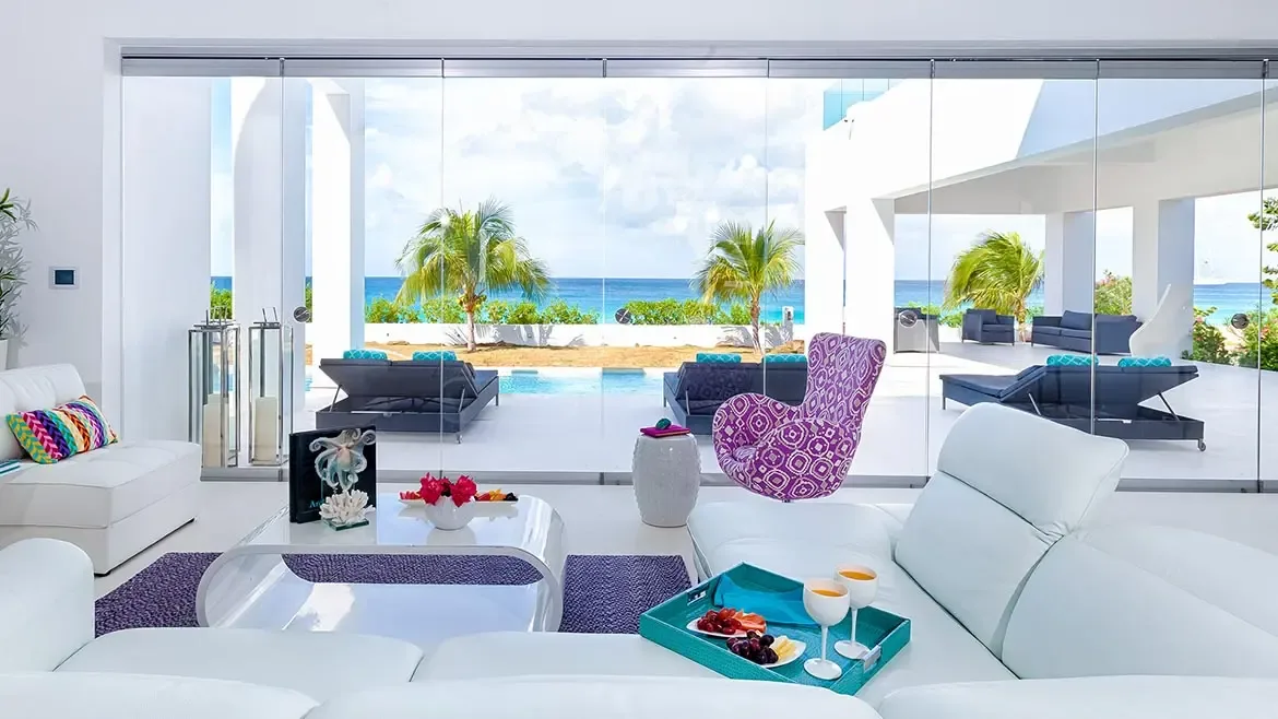 Modern living room with white quilted sofas and chandelier opening to a pool area with red floral trees.