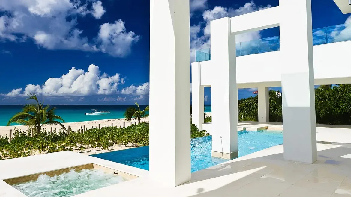 Luxury living room with white quilted chairs and glass table leading to an outdoor pool and tropical view.