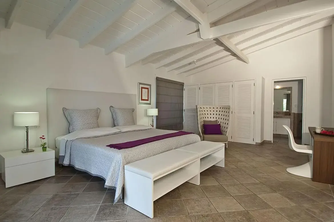 A light-grey bed with a purple runner sits under a vaulted ceiling beside a white wicker chair, where louvered closet doors and a desk with a white S-chair complete the room.