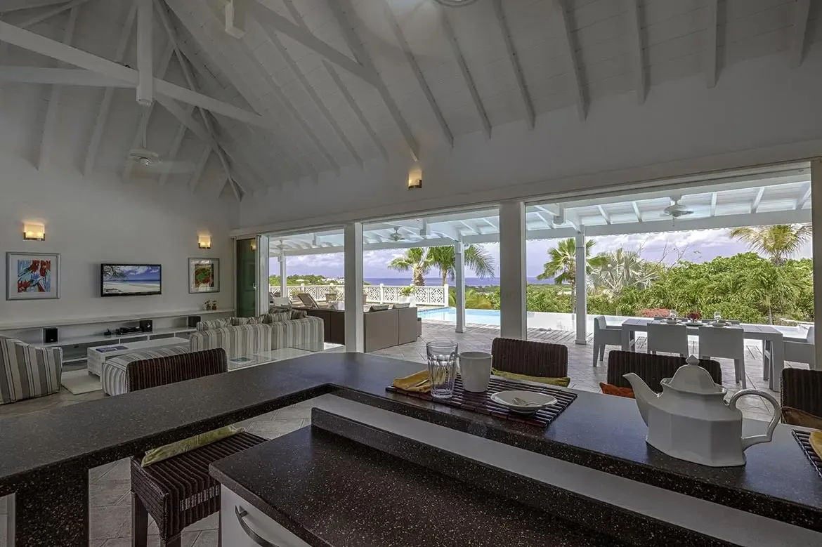 A dark granite breakfast bar with wicker stools fronts a vaulted living room, where open glass doors lead past a white dining table to a turquoise infinity pool and ocean.