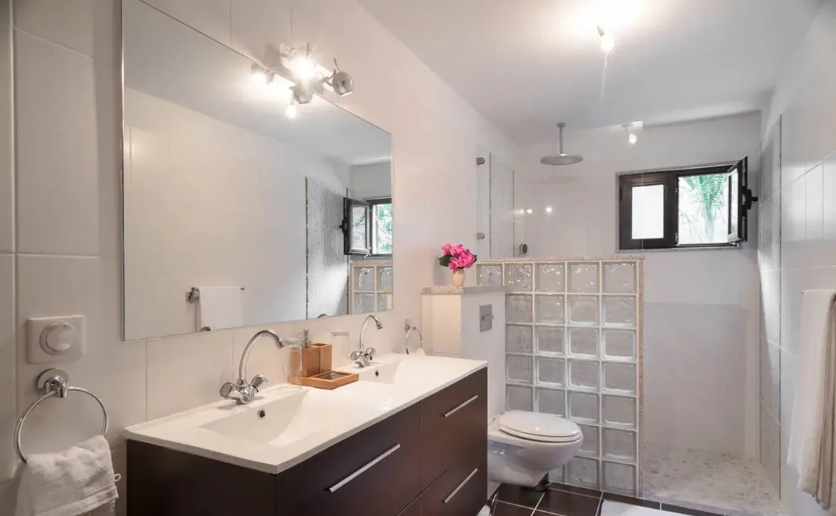 A double vanity with chrome faucets and a glass-block shower partition fill this modern bathroom, where pink flowers and a pebbled shower floor add tropical warmth.