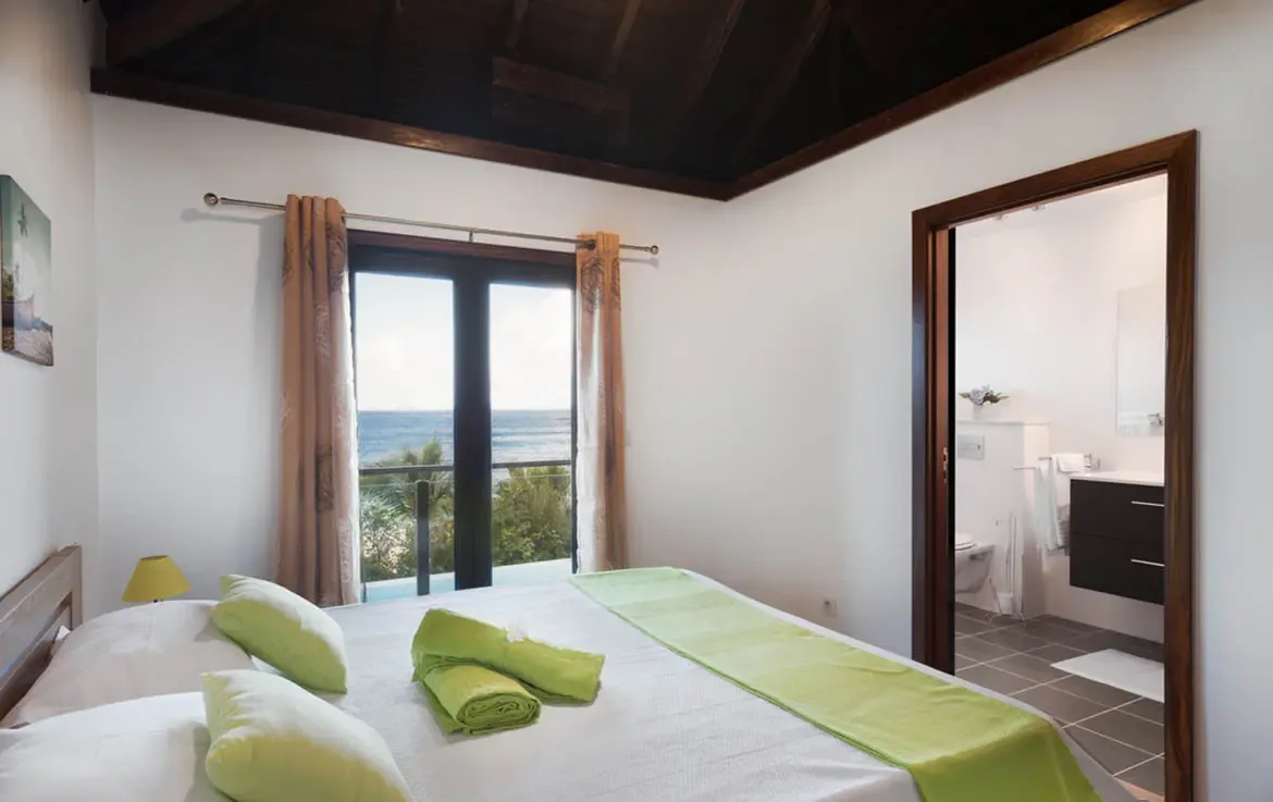 Lime-green pillows and a matching runner accent white linens under a dark vaulted ceiling, where open doors reveal palm trees and a brilliant blue ocean beyond.