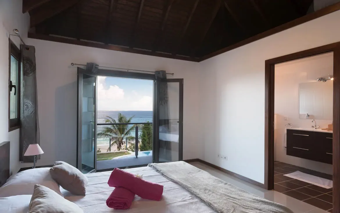 Crisp white linens with rolled pink towels and a tropical flower sit under a vaulted dark-beamed ceiling, where open French doors lead to a balcony overlooking the turquoise ocean.