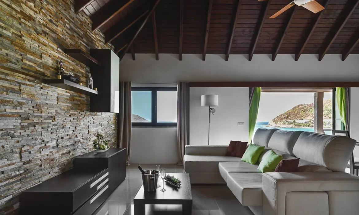 A light leather sectional with lime-green and burgundy pillows faces a stacked-stone accent wall, where open doors framed by green drapes reveal turquoise water and a sandy cove.