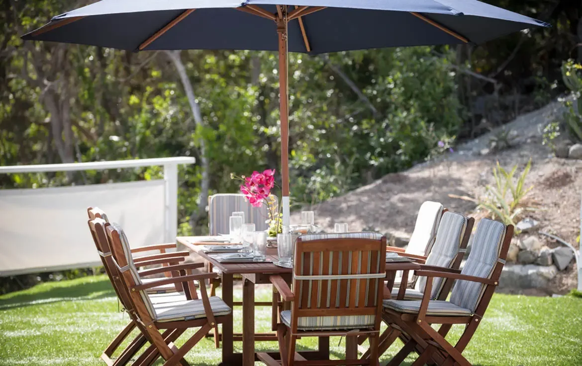 A shaded dining table with striped cushions and a vase of pink orchids sits on a green lawn, where tropical foliage and a white railing enclose a private alfresco setting.