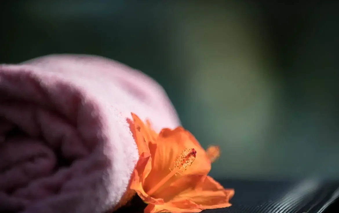 A soft pink rolled towel and a vibrant orange hibiscus bloom rest in dappled shade, where a dark bokeh background evokes a tropical garden hideaway.
