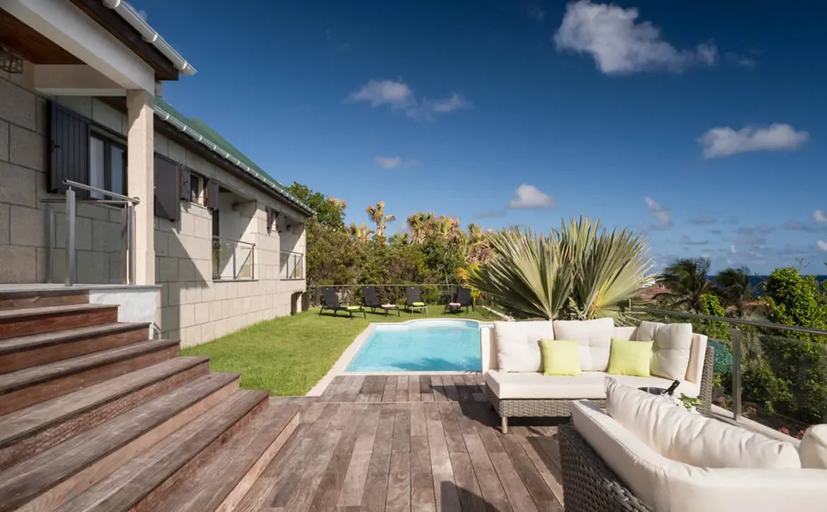 Wooden stairs lead down to a deck with grey wicker sofas and lime-green pillows, where a turquoise pool and black loungers face a deep-blue ocean under scattered clouds.