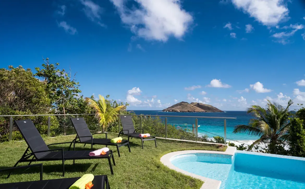 Four black loungers draped with pink and lime-green towels line a poolside lawn, where a glass railing opens to a turquoise bay and a mountainous island on the horizon.
