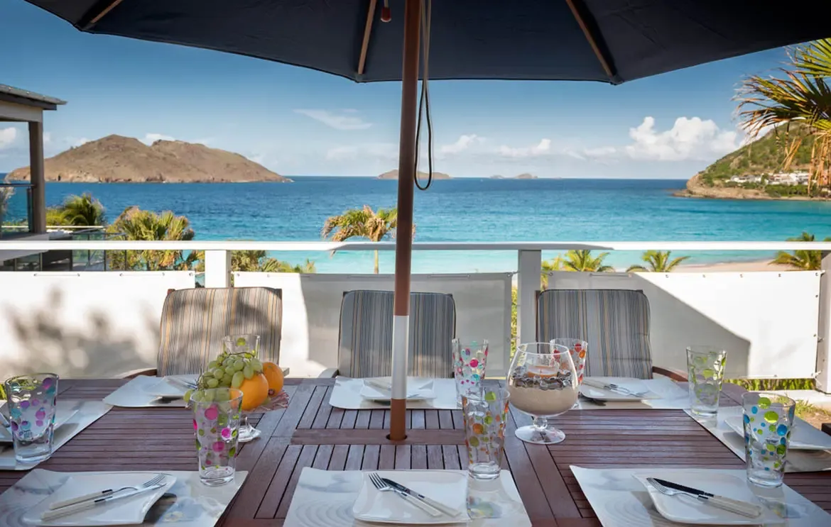 A shaded dining table under a navy umbrella overlooks turquoise bay water and hilly islands, where polka-dot glasses and a fruit bowl await a seaside feast.