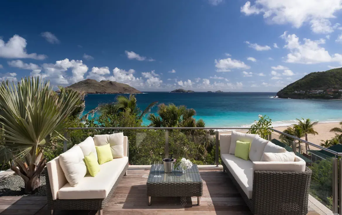 Grey wicker sofas with lime-green pillows and a champagne bucket face a glass-railed terrace, where a fan palm and a crescent beach frame turquoise water stretching to green islands.