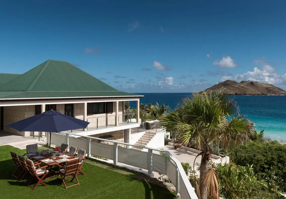 A multi-level stone villa with white pillars and green roof terraces the hillside, where a navy-shaded dining table and loungers face a turquoise bay and a hilly island.