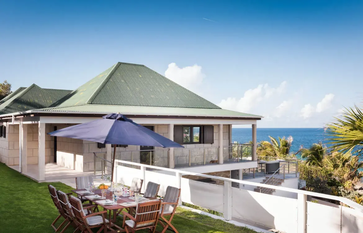 A stone villa with a green corrugated roof and glass-railed balconies overlooks the sea, where a dining table under a navy umbrella sits on a lush lawn.