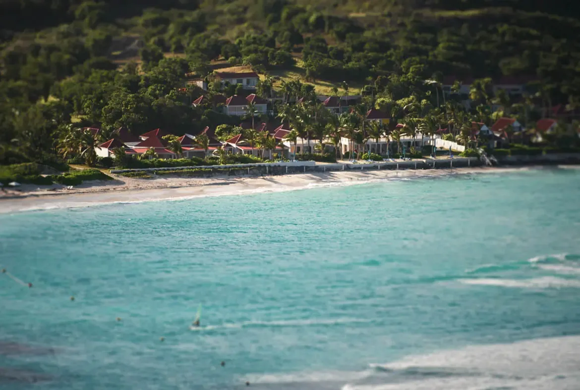 White villas with red gabled roofs sit along a white sand beach, where turquoise water laps the shore and lush green palm trees surround the property at the base of a tropical hillside.