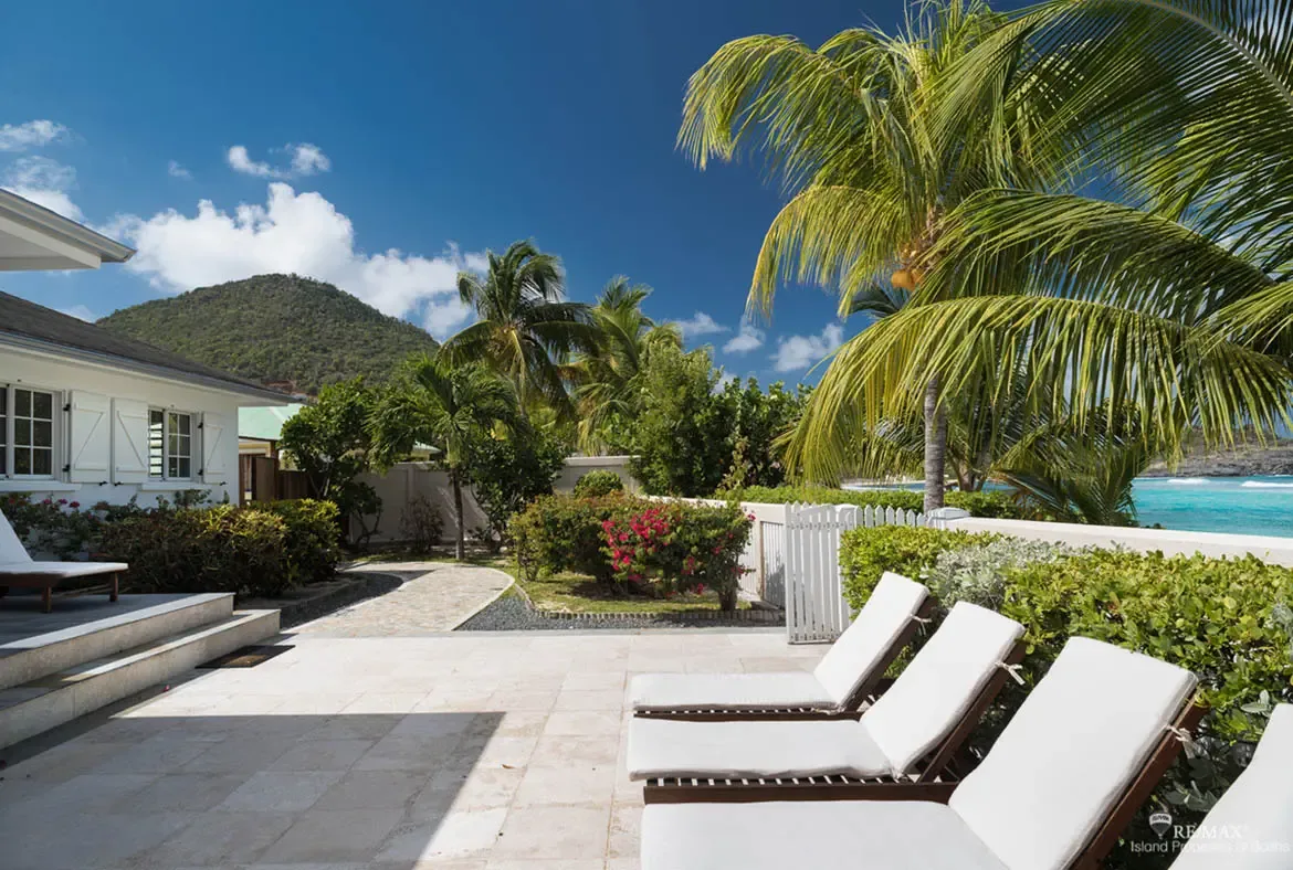 White cushioned loungers sit on a stone patio beside a white picket fence, where pink bougainvillea, coconut palms, and a winding path lead to turquoise ocean glimpses under a blue sky.