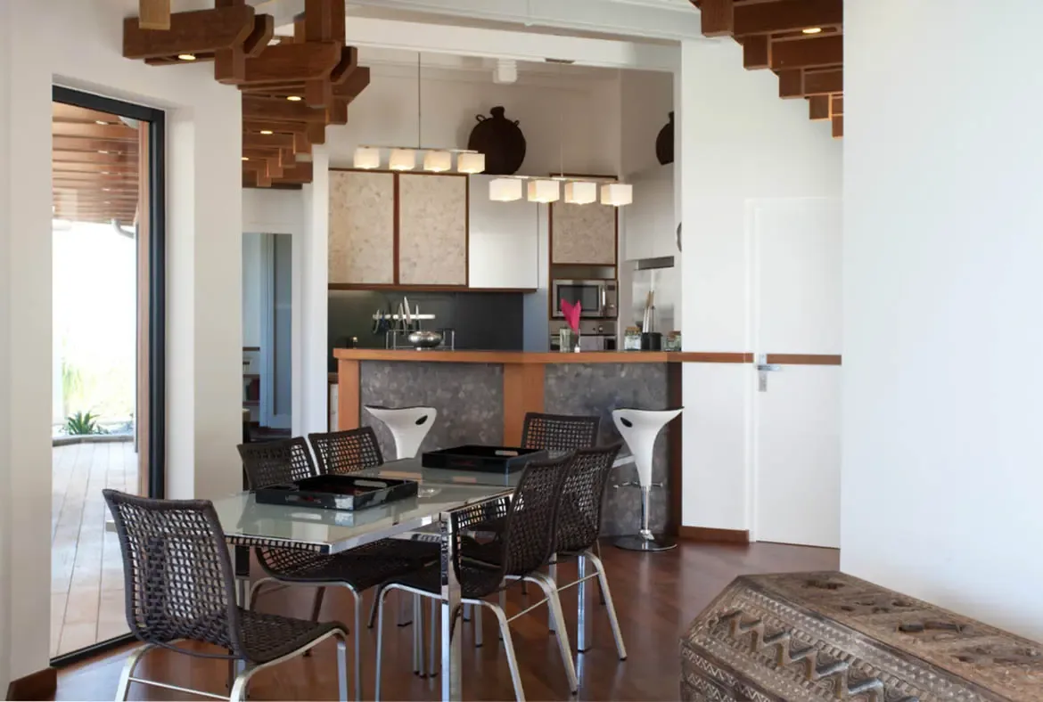 Woven dining chairs and a glass table face a river-stone breakfast bar with white stools, where textured cabinets and sculptural ceiling beams connect to a sunlit deck.