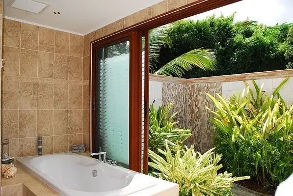 The indoor bathtub sits beside a sliding glass door opening to a private, lushly landscaped outdoor shower area for a tropical bathing experience.