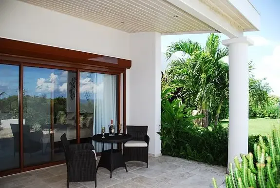 A covered outdoor patio features wicker seating and a small dining table with views of lush tropical landscaping through large glass sliding doors.