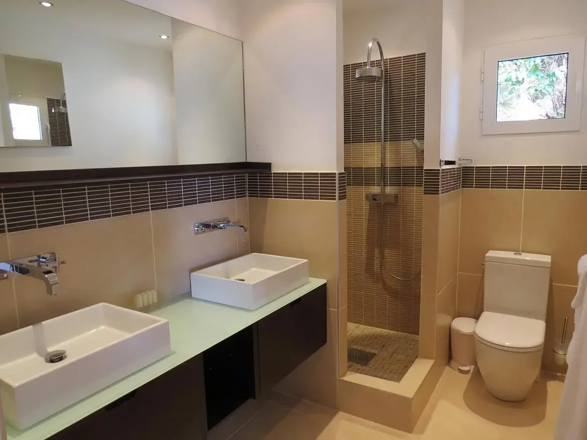 A modern bathroom featuring double vanity with two white vessel sinks, wall-mounted faucets, large mirror, walk-in shower with dark mosaic tile accents, and contemporary white toilet.