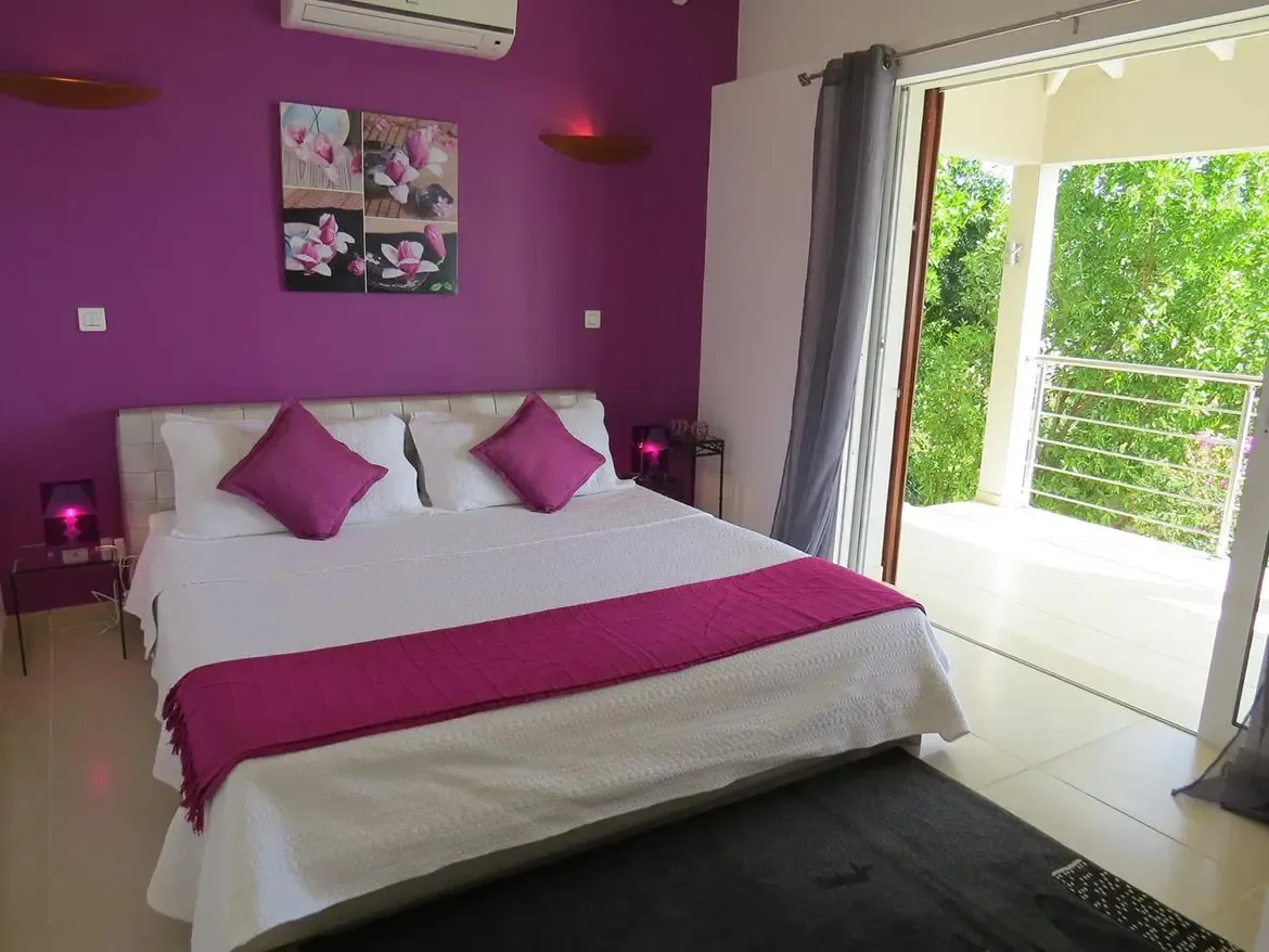 A modern bedroom with vibrant purple accent wall, white bedding with purple runner, wall-mounted art, and open flow to a private balcony with lush greenery views.