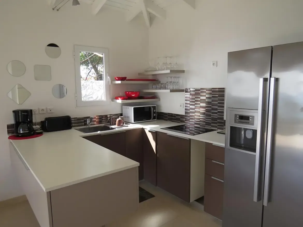 A modern U-shaped kitchen featuring white countertops, brown cabinetry, patterned tile backsplash, stainless steel refrigerator, microwave, and open shelving with red dishware.